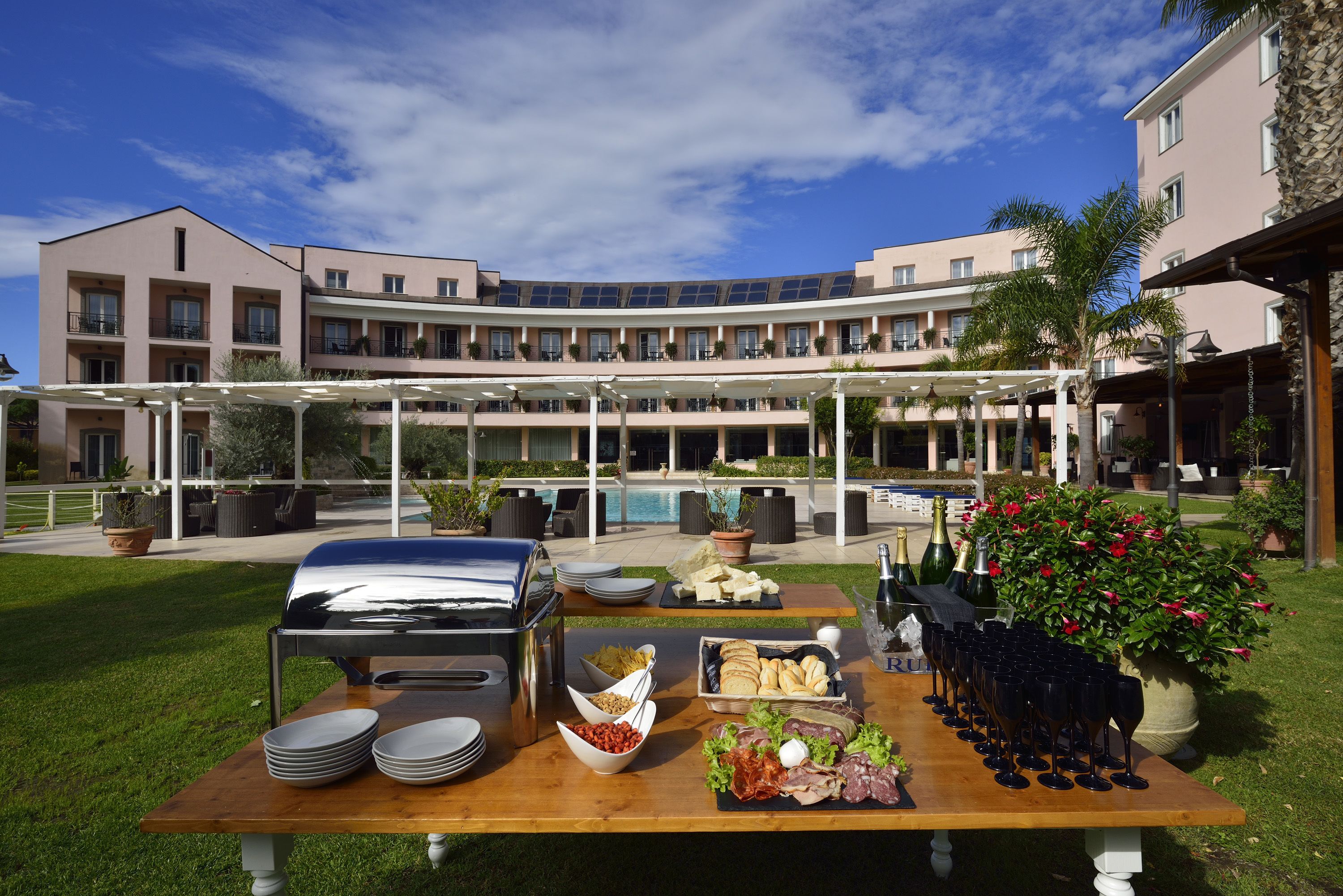 An outdoor dining setup with a barbecue grill, plates, bowls of food, and wine glasses, set against a backdrop of a multi-story building with balconies and a pool.