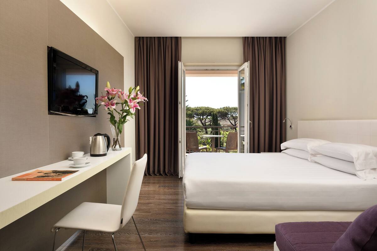Habitación de hotel con cama, escritorio con silla, televisor montado en la pared y ventana con cortinas.