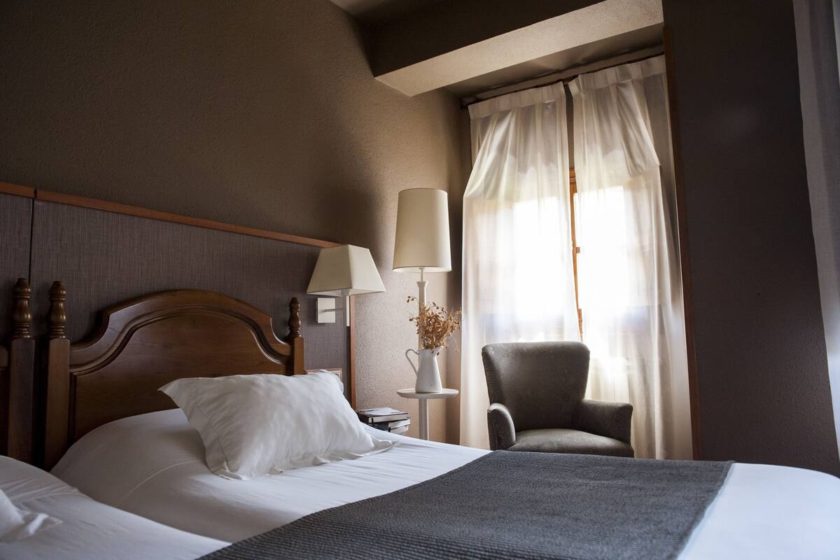 Une chambre d’hôtel avec une tête de lit en bois, un lit recouvert de draps blancs, un plaid gris, une table de chevet avec une lampe et un vase, et une chaise près d’une fenêtre ornée de rideaux légers.