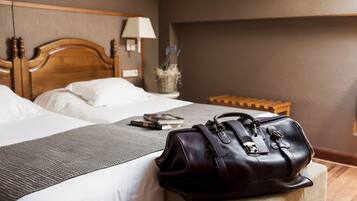 Habitación de hotel con dos camas, una mesita de noche con lámpara, una planta pequeña y un bolso de cuero sobre un taburete.