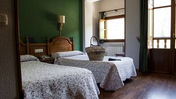 A room with two single beds, a wooden headboard, a bedside table with a lamp, a window with curtains, and a radiator.