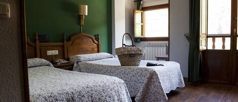 Una habitación con dos camas individuales, un cabecero de madera, una mesita de noche con lámpara, una ventana con cortinas y un radiador.