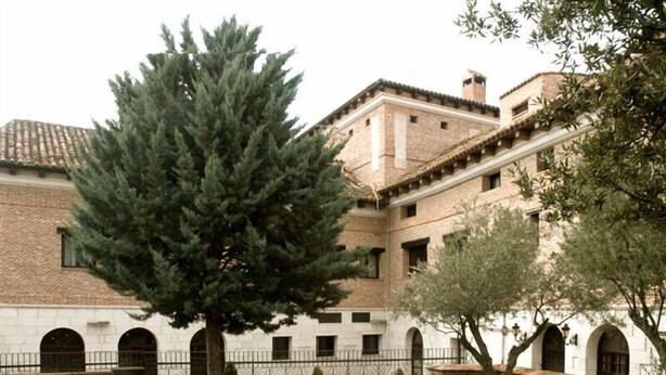 A courtyard with a large evergreen tree, a white building with arched windows, and a brick wall. - Hotel El Jardín de la Abadía (Arroyo de la Encomienda)
