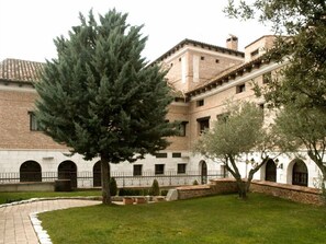 A courtyard with a large evergreen tree, a white building with arched windows, and a brick wall. - Hotel El Jardín de la Abadía (Arroyo de la Encomienda)