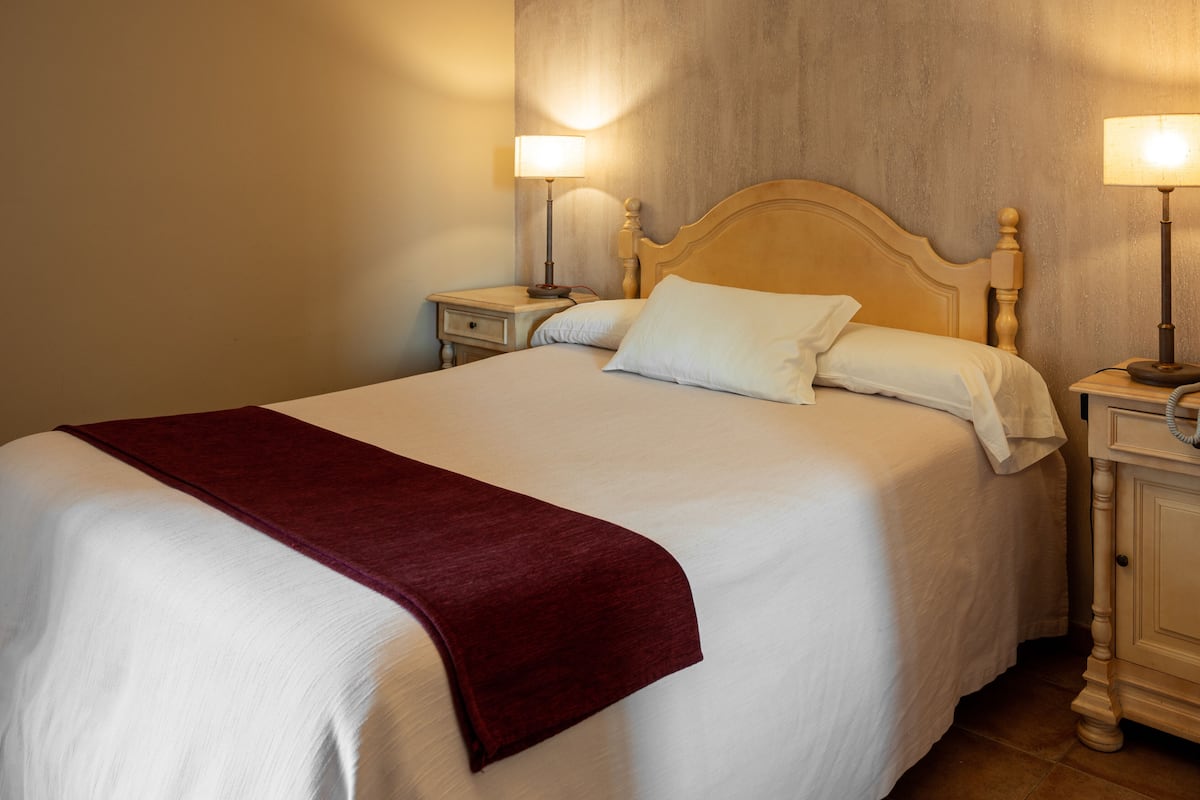Un lit bien fait, recouvert d’une couette blanche et agrémenté d’un plaid rouge, flanqué de deux tables de chevet avec des lampes, dans une pièce dotée d’une tête de lit en bois et d’un mur à texture.