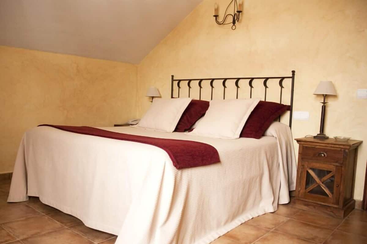 Un lit simple avec un cadre en métal, une literie blanche et un plaid rouge. Il y a deux lampes de chevet et une table de chevet en bois avec un plateau en verre. La pièce a un plafond en pente et un sol carrelé.