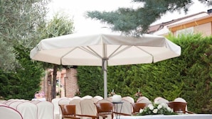 An outdoor seating area with white tablecloths and chairs, a large umbrella, and a floral centerpiece. - Hotel El Jardín de la Abadía (Arroyo de la Encomienda)