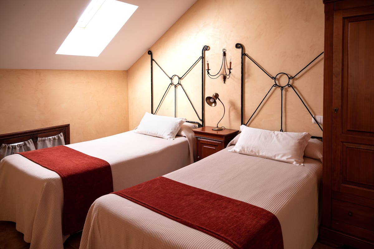 Deux lits simples avec du linge de lit blanc et des plaids rouges, une table de chevet en bois, une lampe fixée au mur et une armoire en bois.