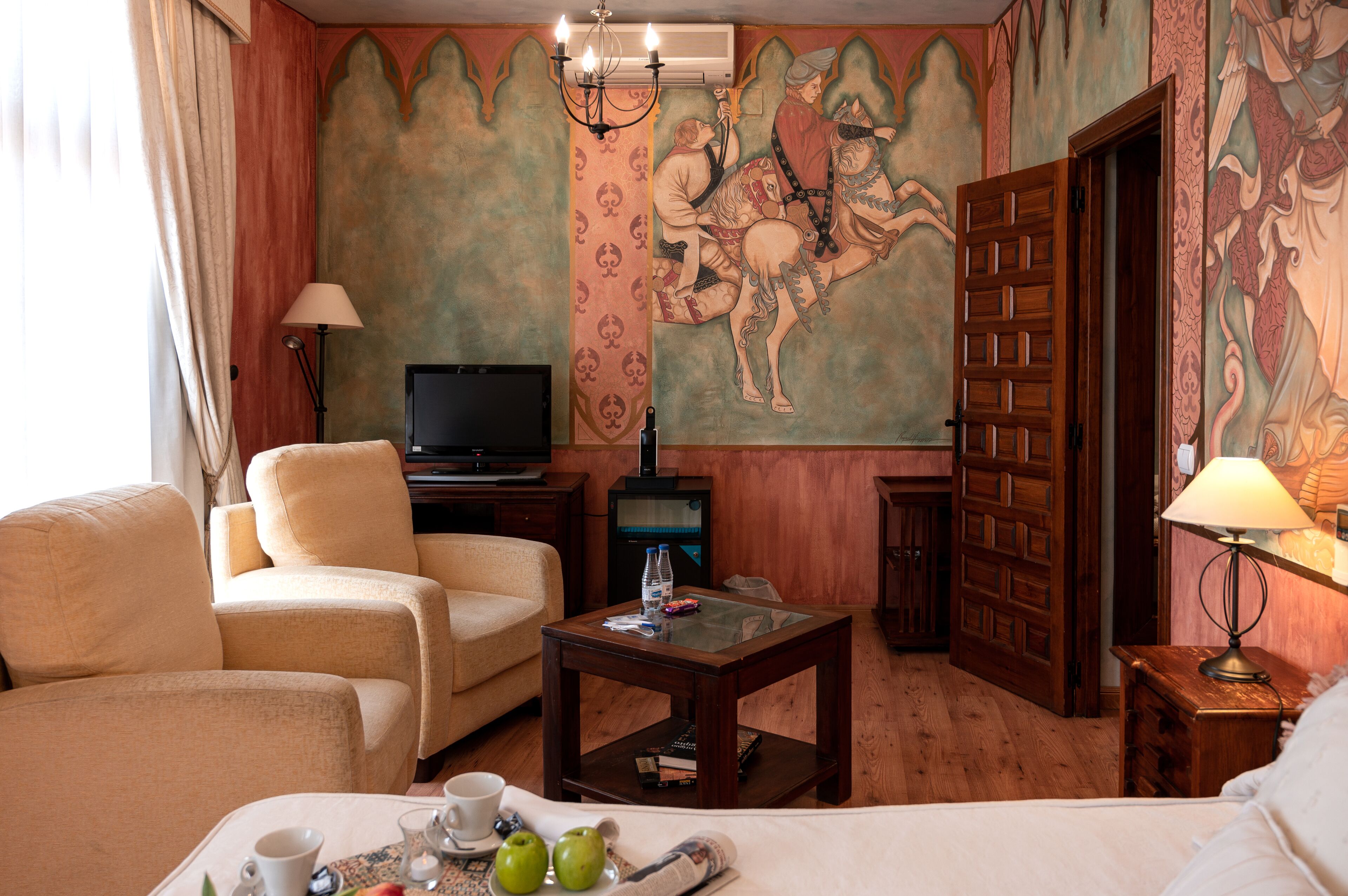 A hotel room with a large bed, two armchairs, a wooden coffee table, a television, and a mural on the wall.