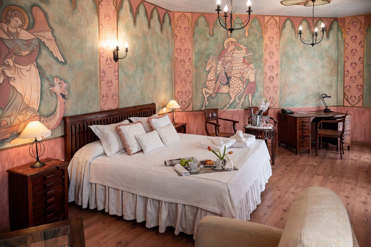 Une chambre spacieuse avec un grand lit, un bureau et une chaise. Les murs sont décorés de fresques et la pièce est agrémentée d’un lustre.