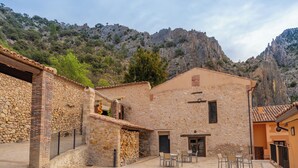 Un edificio de piedra con patio, mesas al aire libre y montañas al fondo.