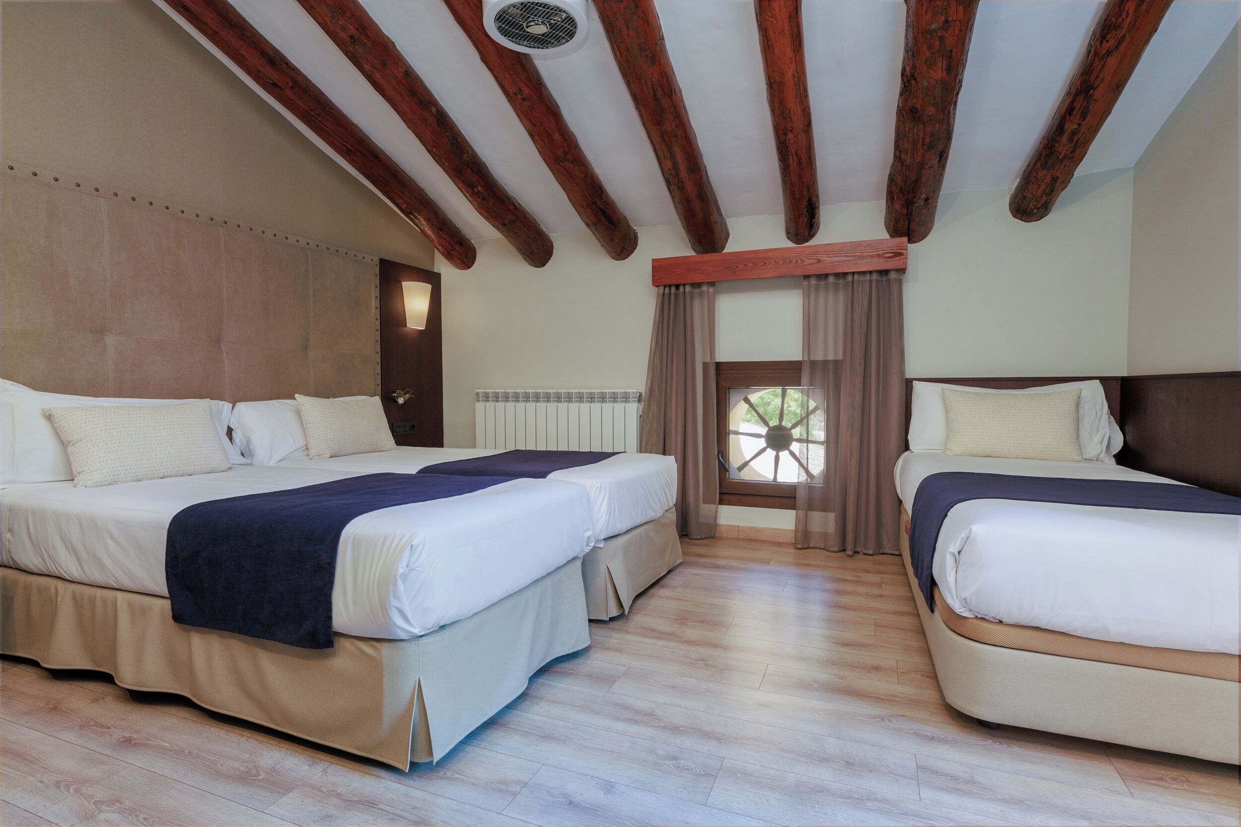 Habitación de hotel con dos camas, vigas de madera en el techo, una ventana con cortinas y un radiador.