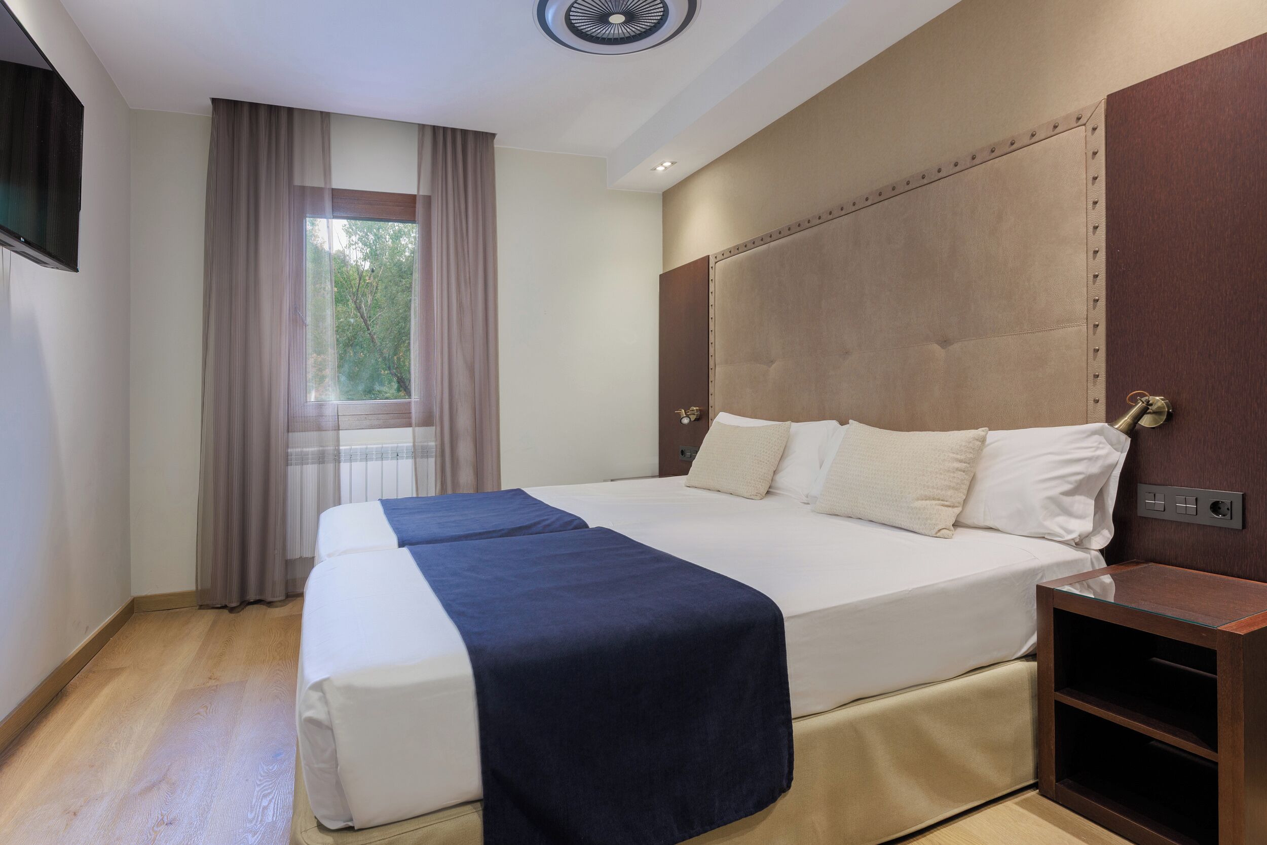 Ein Hotelzimmer mit einem großen Bett, einem Nachttisch, einem Fenster mit Vorhängen und einem Deckenventilator.
