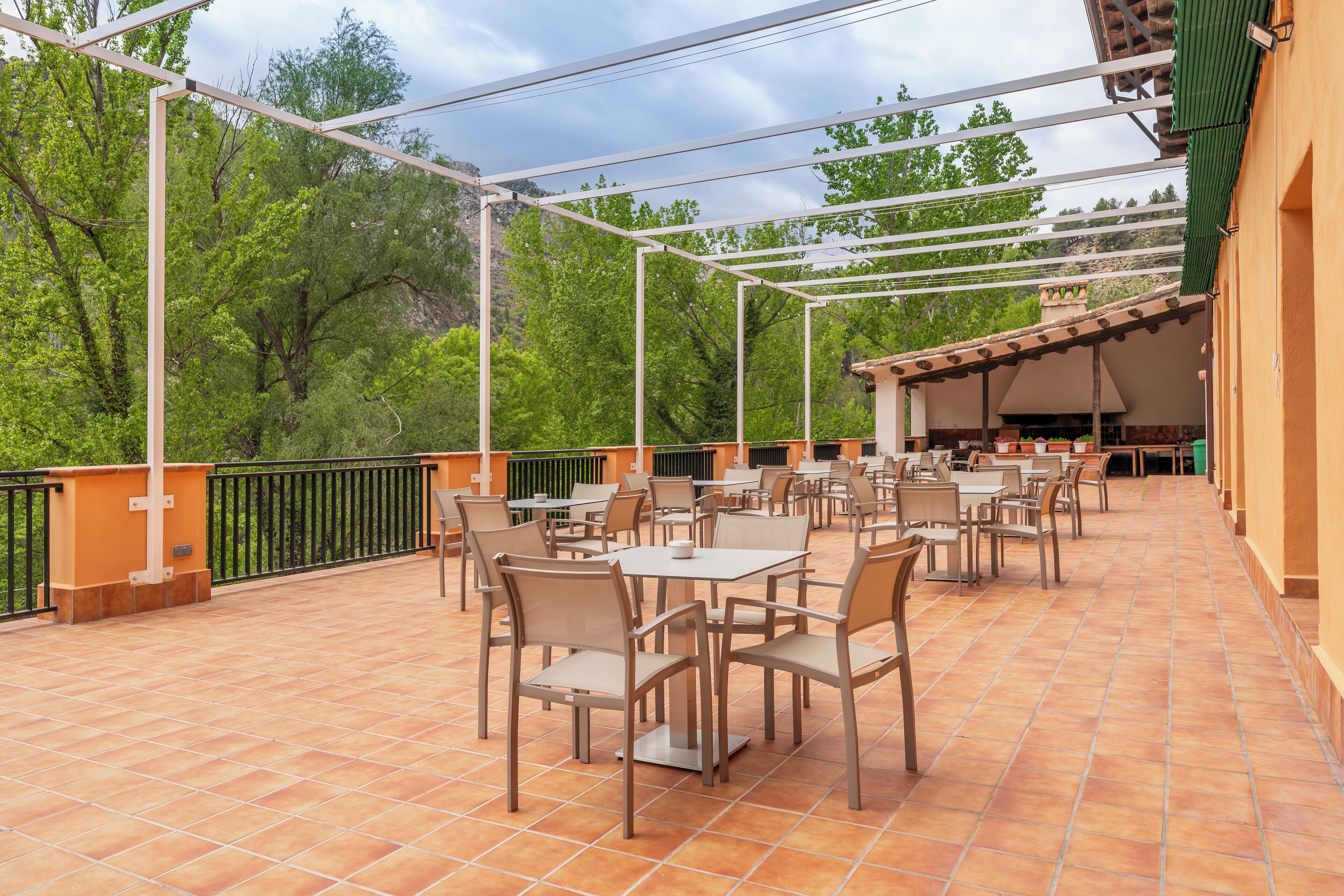 Un patio al aire libre con mesas y sillas, rodeado por una barandilla de metal y árboles.