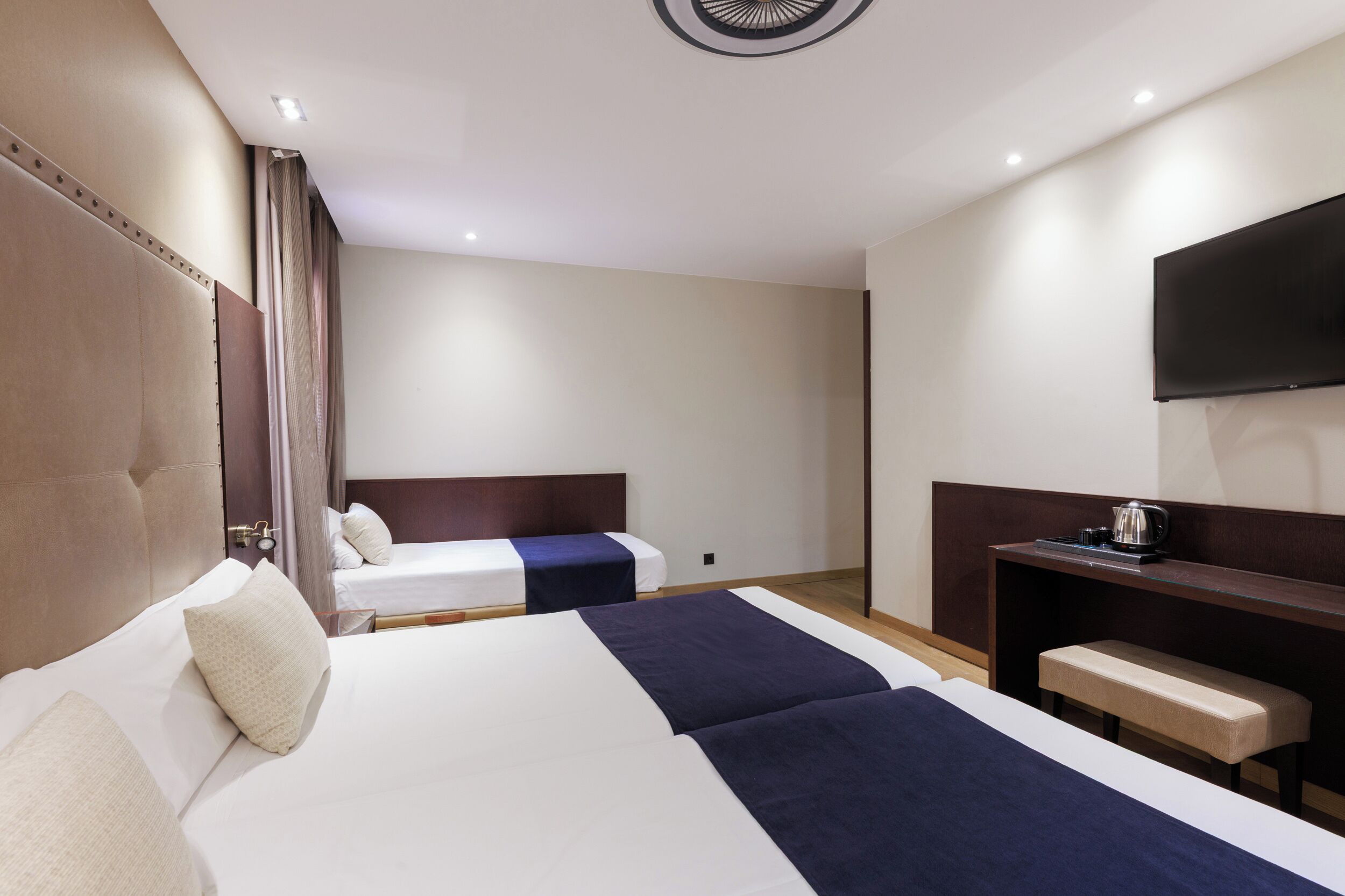 A hotel room with two beds, a desk, a flat-screen TV, and a ceiling light fixture.