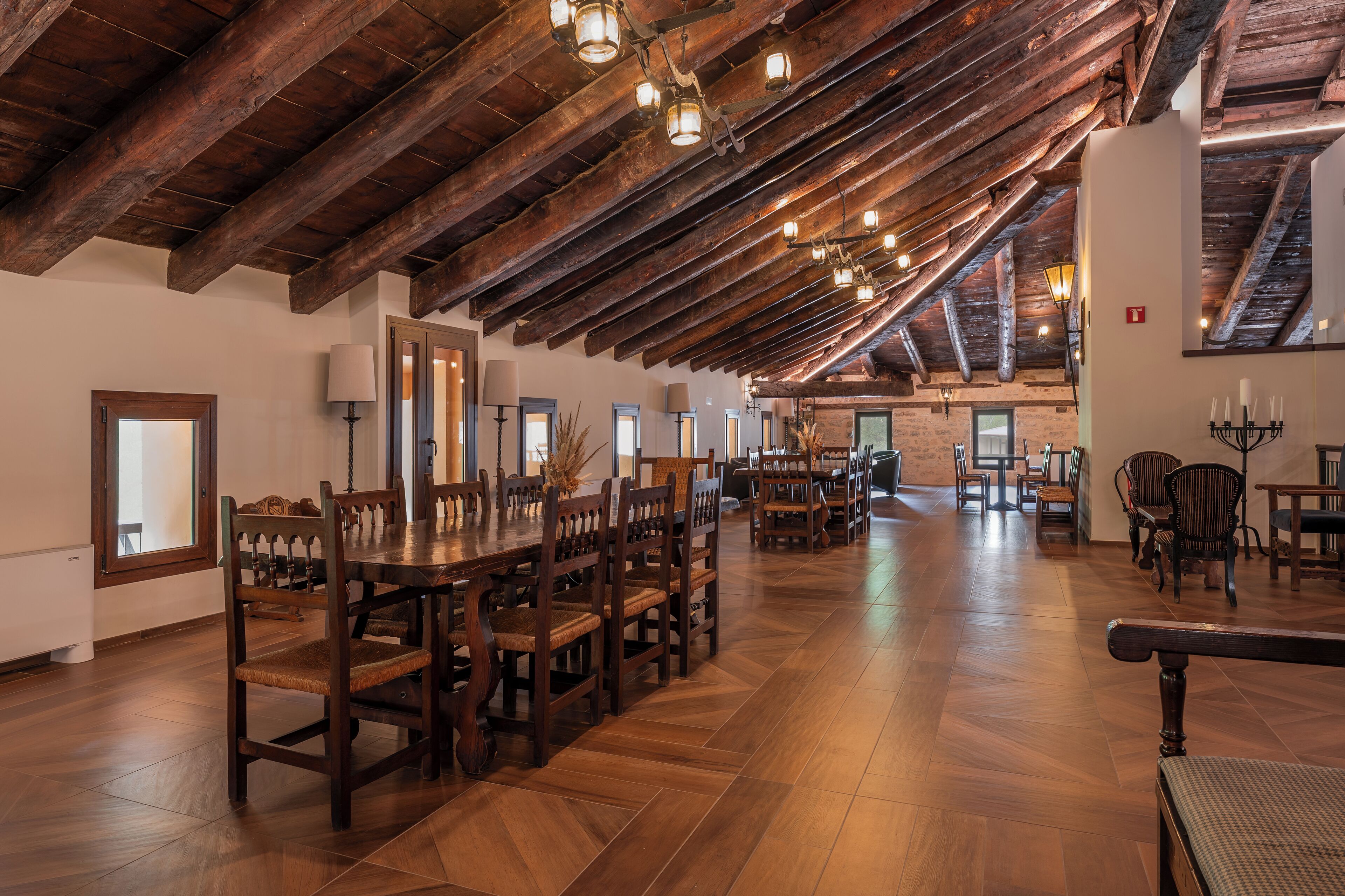 A spacious dining area with wooden tables and chairs, a high wooden ceiling, and a view of the kitchen in the background.