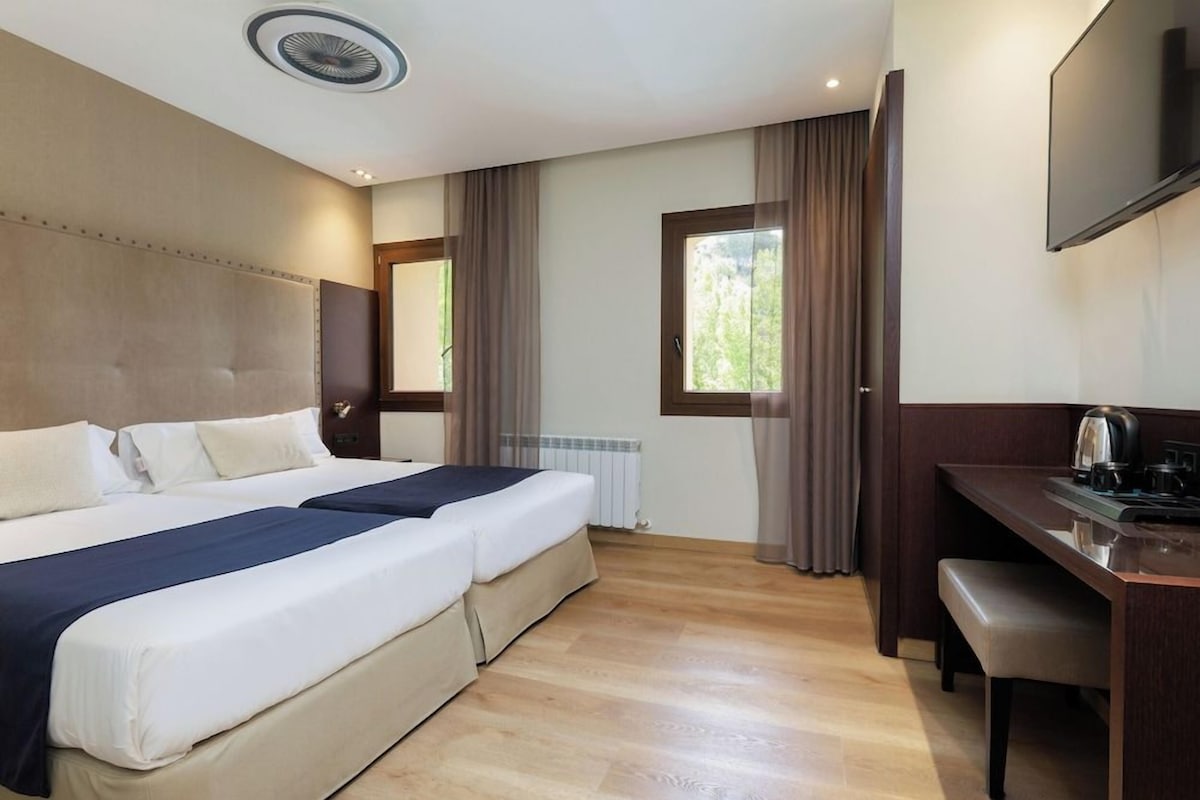 Una habitación de hotel con cama doble, un escritorio con silla, televisión y ventana con cortinas.