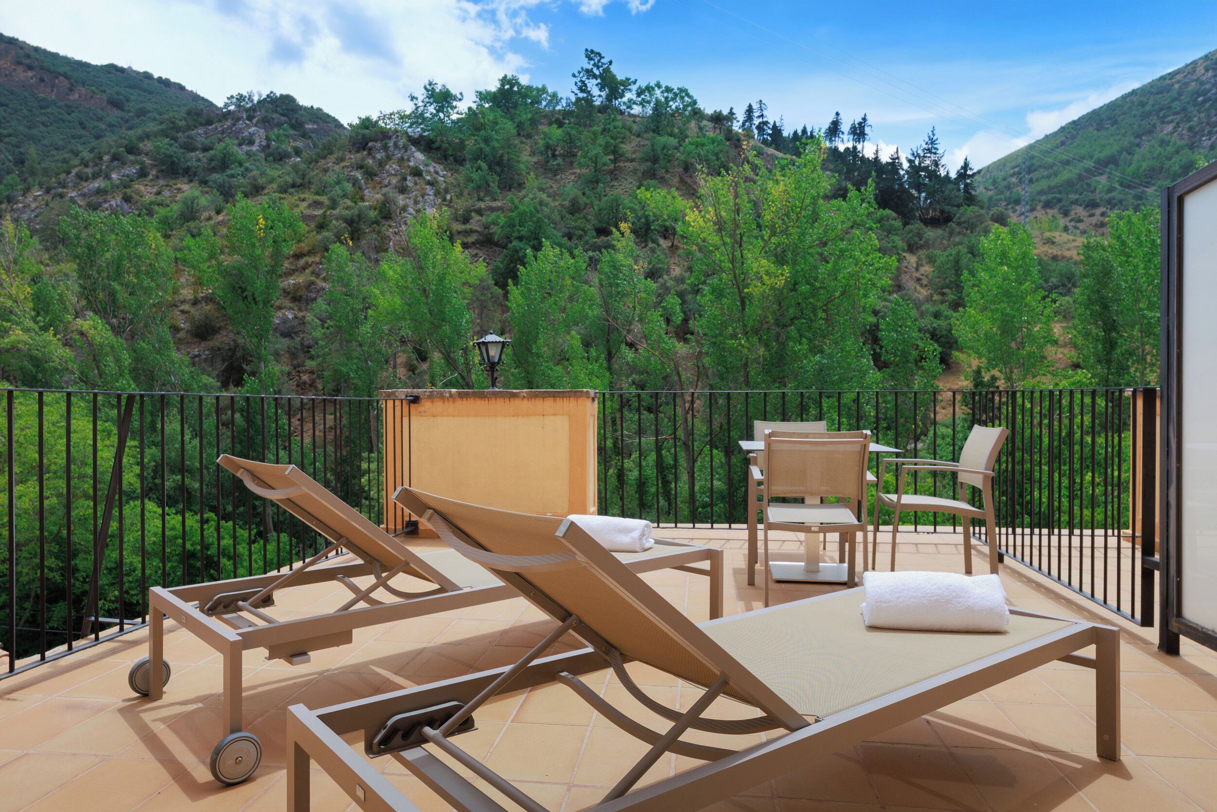 A balcony with sun loungers, a table, and chairs, overlooking a mountainous landscape.