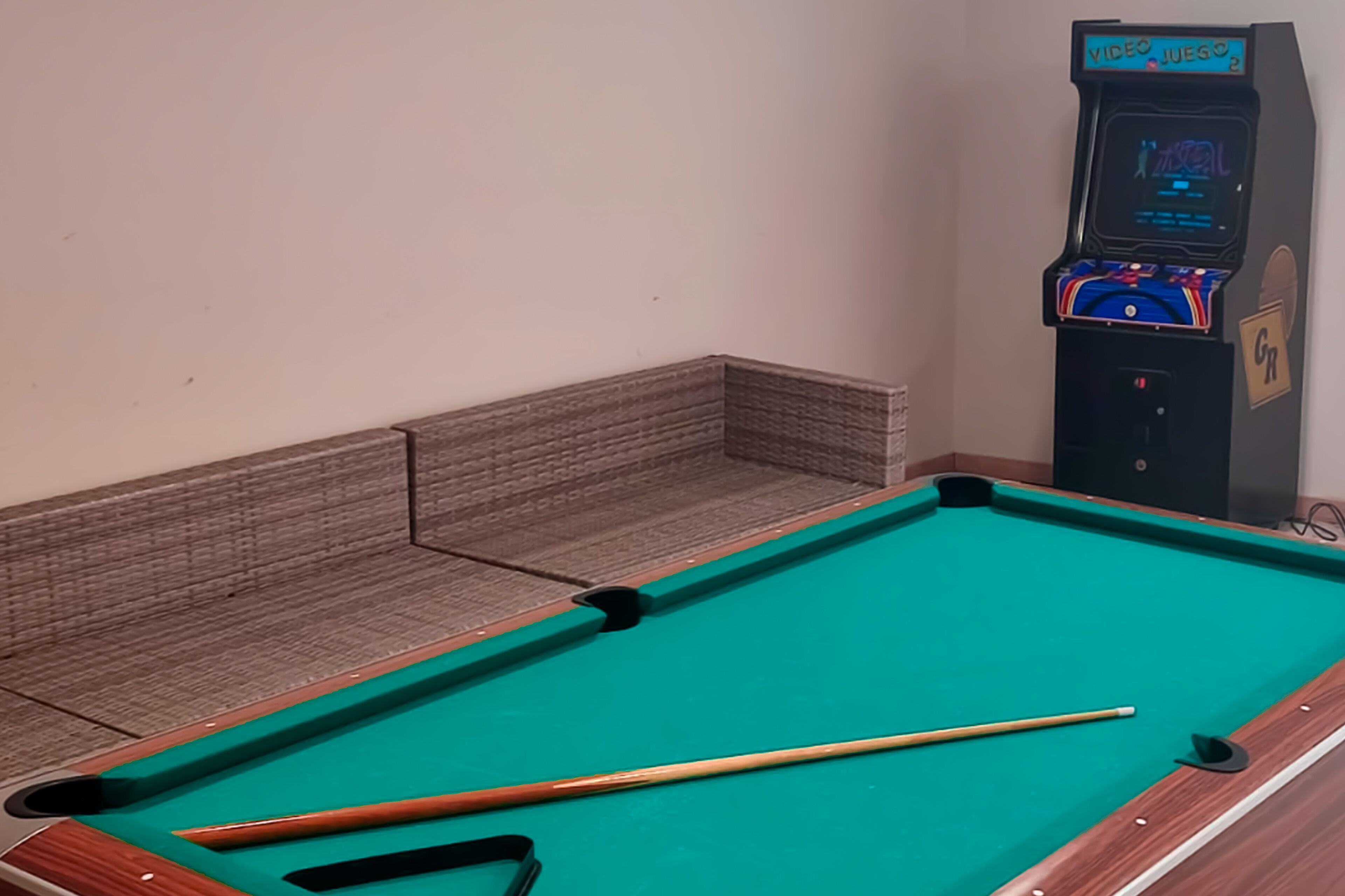 A pool table with a pool cue and a video game machine in the corner.