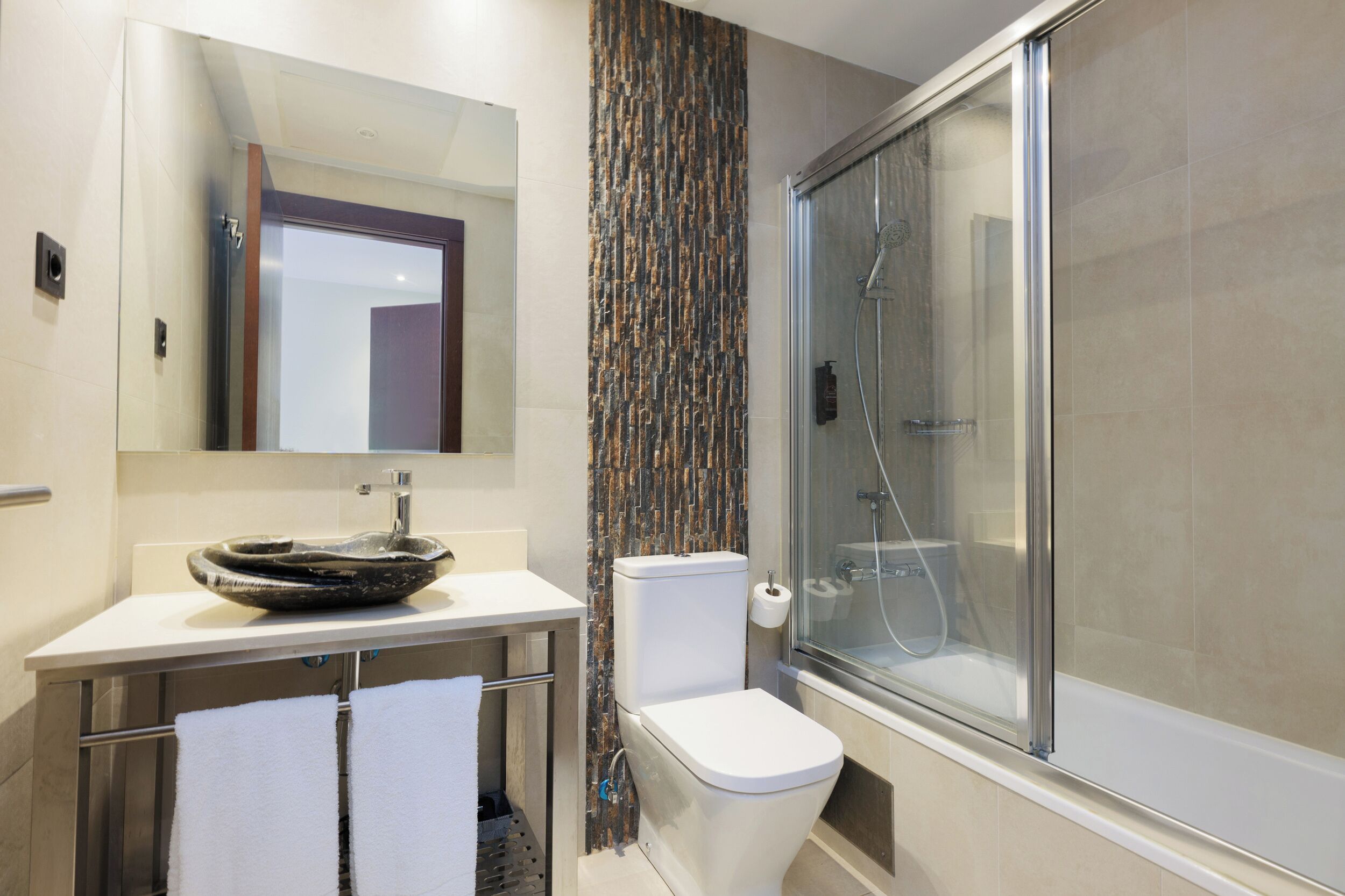 A modern bathroom with a glass shower enclosure, a white sink with a unique basin, and a toilet with a vertical tile backsplash.