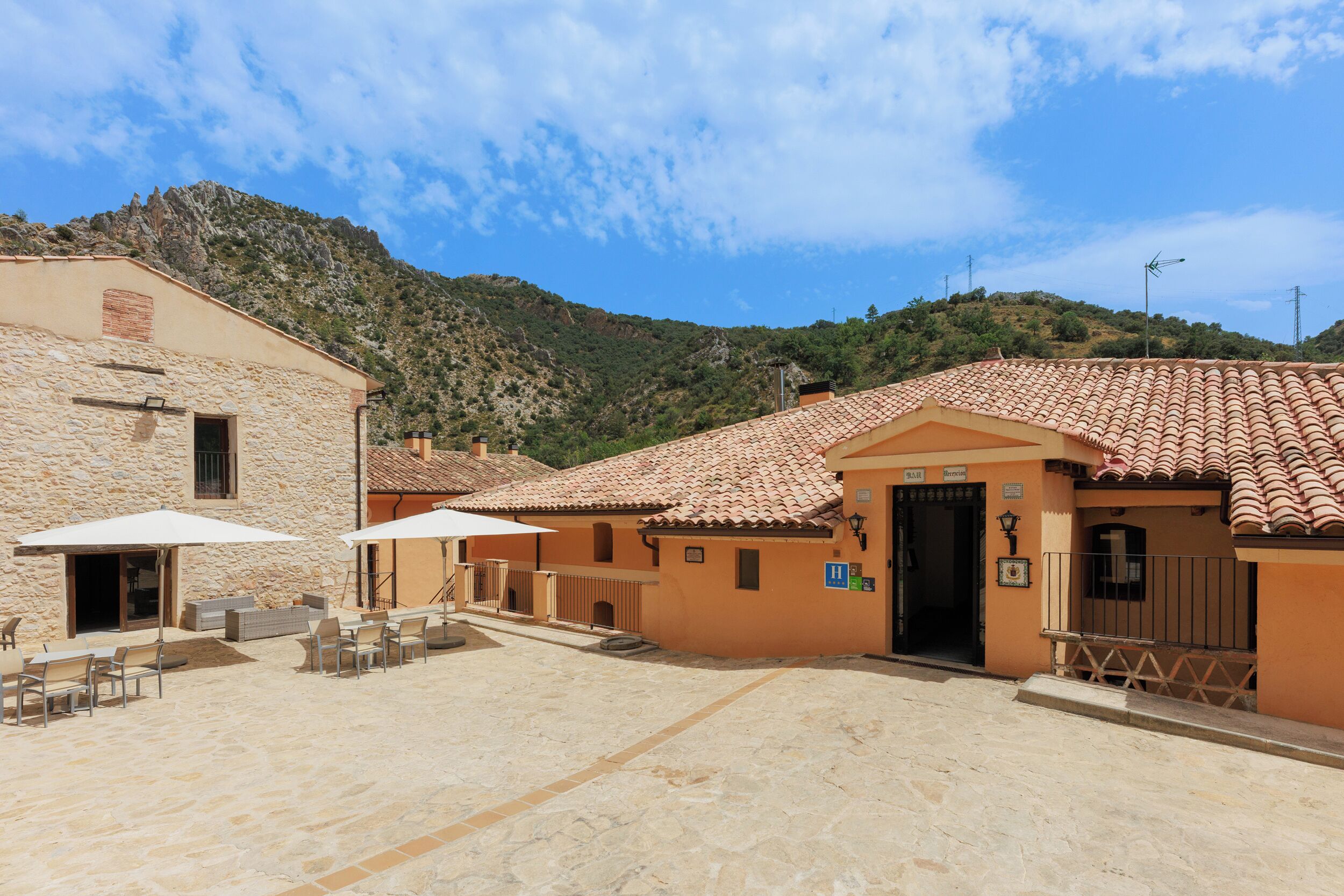 Un patio con edificios de piedra, mobiliario exterior y un paisaje montañoso al fondo.