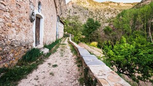 Un estrecho sendero de piedra que conduce a un edificio rústico con puerta de madera, rodeado de vegetación y montañas al fondo.