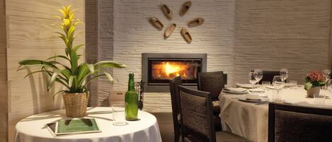 A dining area with a fireplace, a table set for two, and a potted plant.