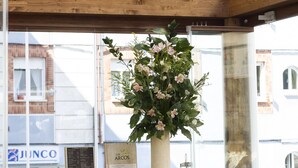 A restaurant interior with wooden beams, a hanging pendant light, wicker chairs, a large floral centerpiece, and a menu board.