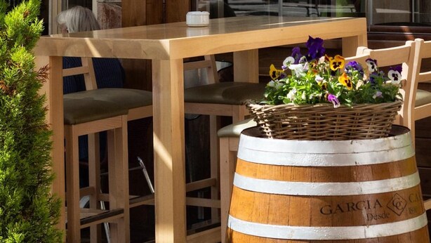 A wooden outdoor seating area with a wooden table and chairs, a wicker basket with flowers, and a wooden barrel with text.