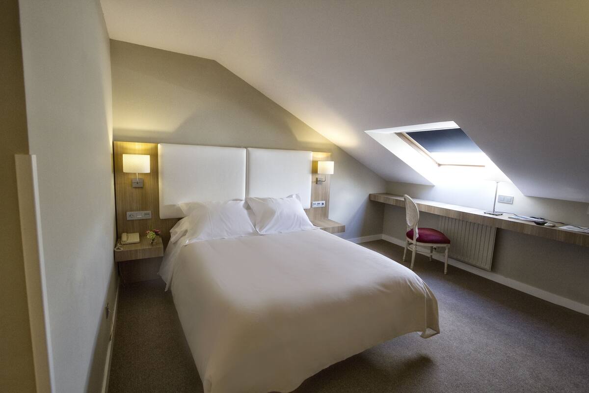 Una habitación de hotel con una cama grande, un escritorio con silla, una mesita pequeña y un tragaluz.