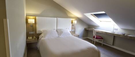 Una habitación de hotel con una cama grande, un escritorio con silla, una mesita pequeña y un tragaluz.