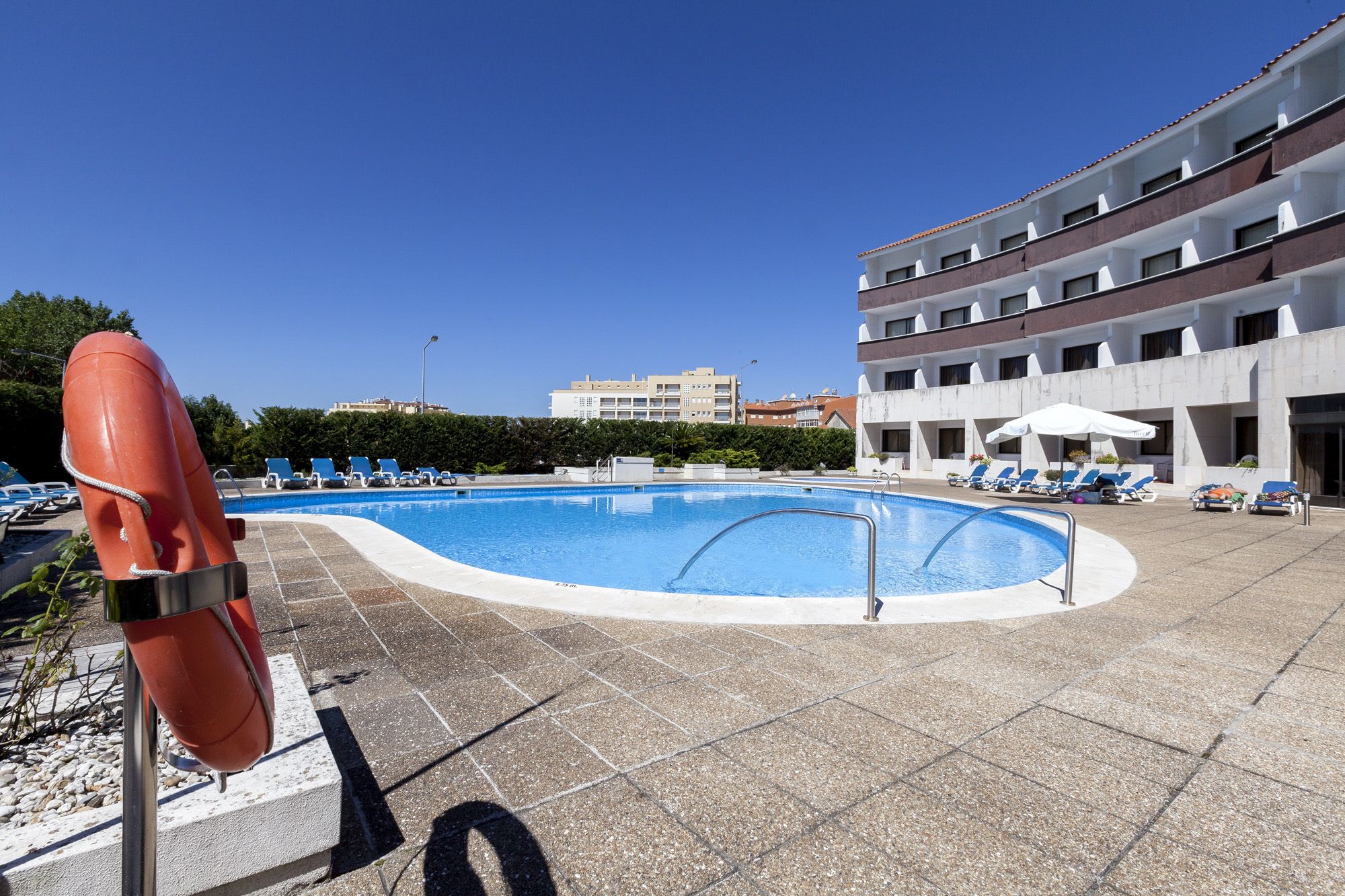 A swimming pool with lounge chairs and umbrellas, a lifebuoy, and a building with multiple balconies.
