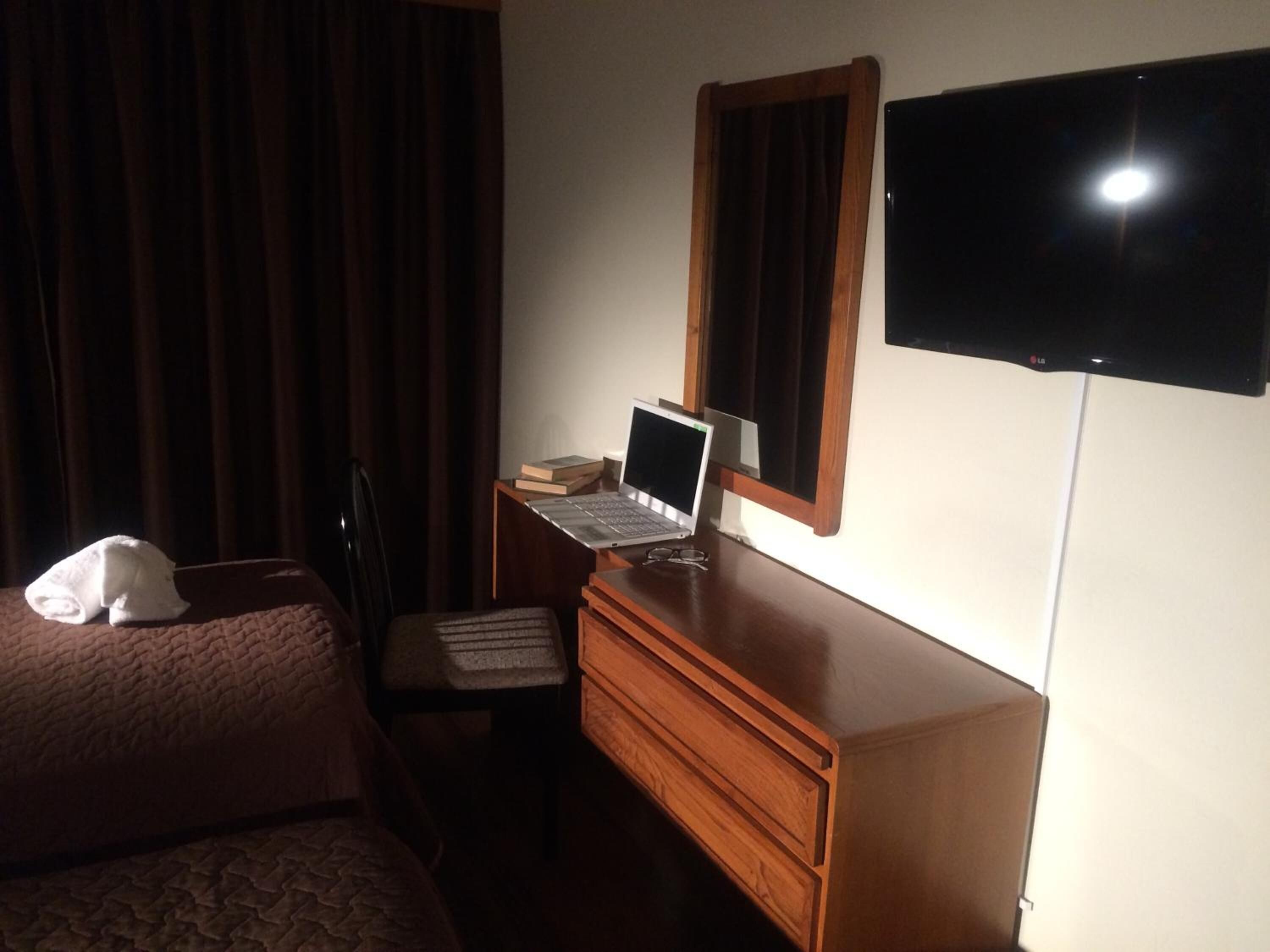 Habitación de hotel con cama, escritorio con una computadora portátil, una silla, televisión y ventana con cortinas.