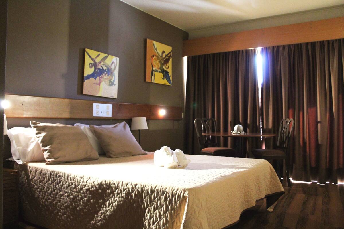 A hotel room with a bed, two pillows, a bedside table with a lamp, a chair, a table with a vase, and two paintings on the wall.