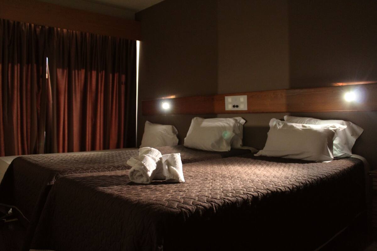 A hotel room with a large bed, two pillows, a headboard with a light, and a window with curtains.