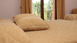 Una cama con un edredón tejido y dos almohadas, una ventana con cortinas y vistas al exterior con vegetación.