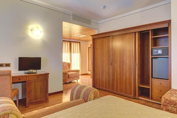 Habitación de hotel con armario de madera, escritorio con televisor, sillón estampado y cama con colcha estampada.