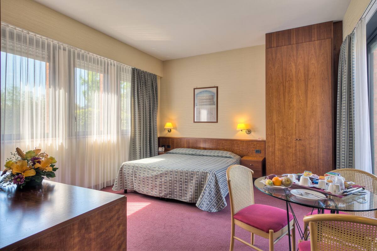 Ein Hotelzimmer mit einem Bett, einem Schreibtisch mit Blumenvase, einem Esstisch mit Frühstücksgebäck und einem großen Fenster mit Vorhängen.