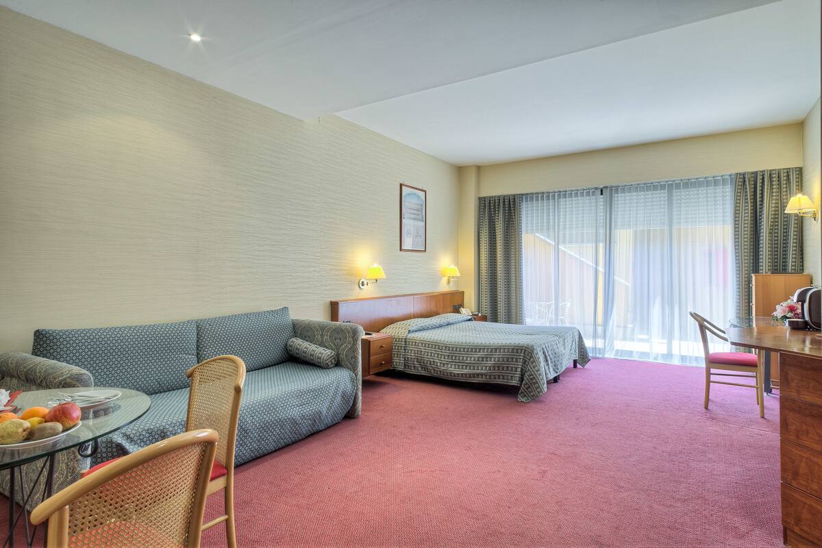 Ein Hotelzimmer mit einer Couch, zwei Einzelbetten, einem Schreibtisch, einem Stuhl und einem kleinen Tisch mit Früchten.