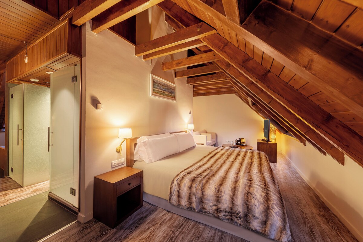 Un dormitorio con techo inclinado, vigas de madera, cama con manta a rayas, lámparas de noche y un cuadro en la pared.