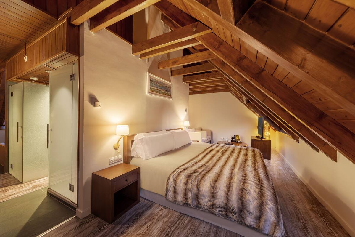 Une chambre avec un plafond en pente, des poutres en bois, un lit recouvert d’une couverture à rayures, des lampes de chevet et un tableau accroché au mur.