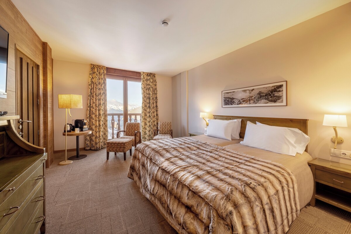 Habitación de hotel con una cama grande, un escritorio con lámpara, una silla, una ventana con cortinas y un cuadro en la pared.
