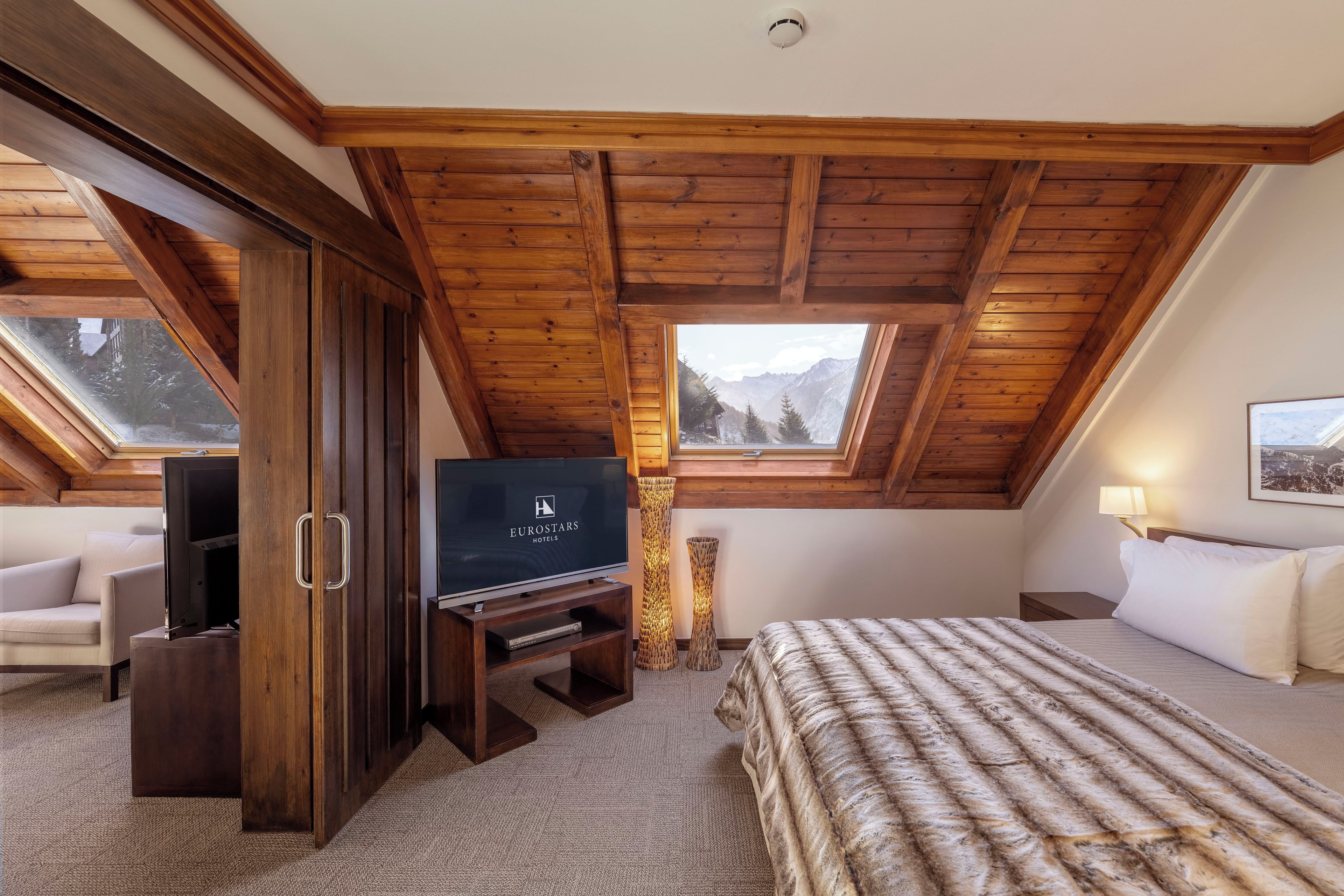 Une chambre d’hôtel avec un plafond en pente, une télévision, un lit, un canapé et une fenêtre donnant sur la montagne.