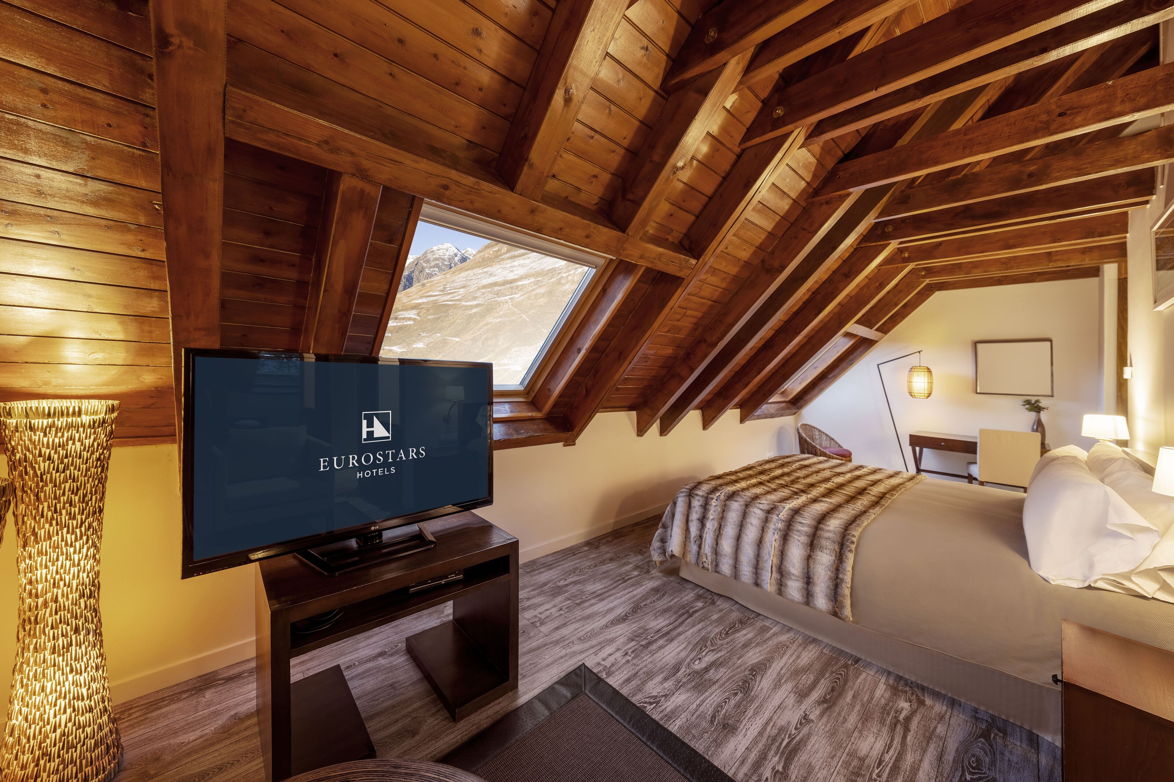 A room with a wooden ceiling, a bed, a TV, and a window with a mountain view.