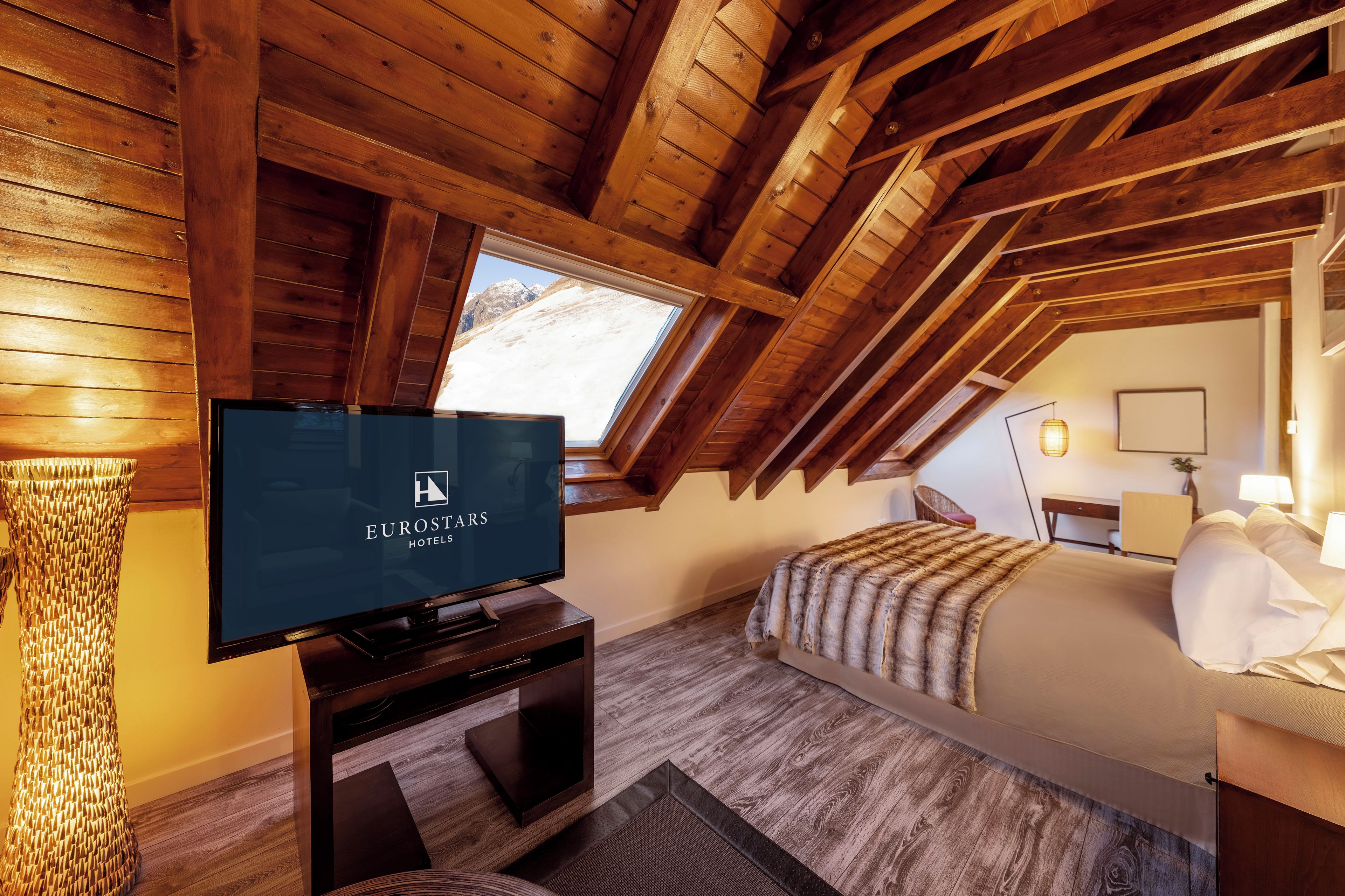 A hotel room with a television displaying 'EUROSTARS HOTELS' on the screen, wooden beams on the ceiling, a bed with a fur throw, and a window overlooking a snowy landscape.
