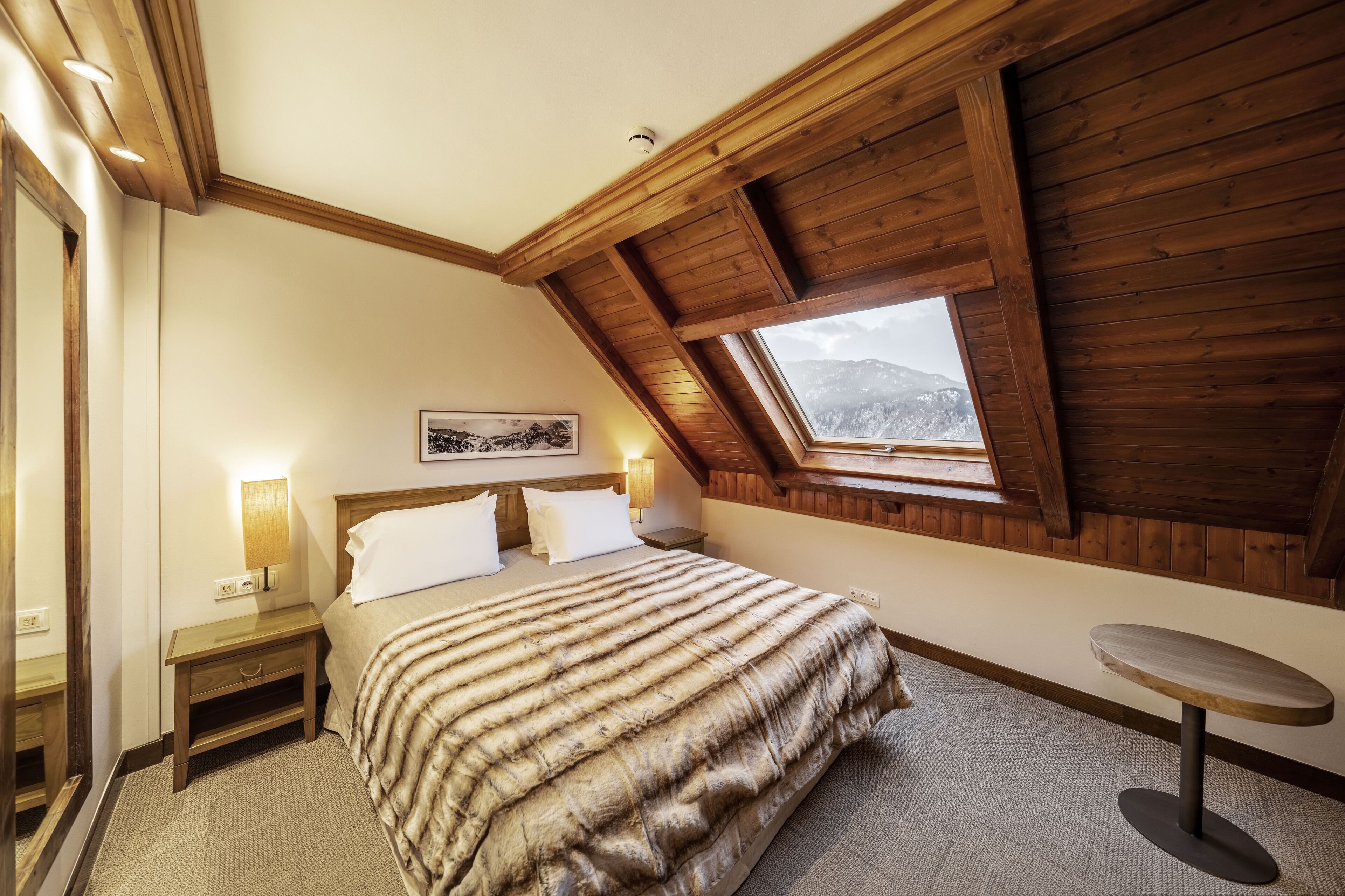 A bedroom with a sloped ceiling, a bed, a nightstand, a table, and a window with a mountain view.