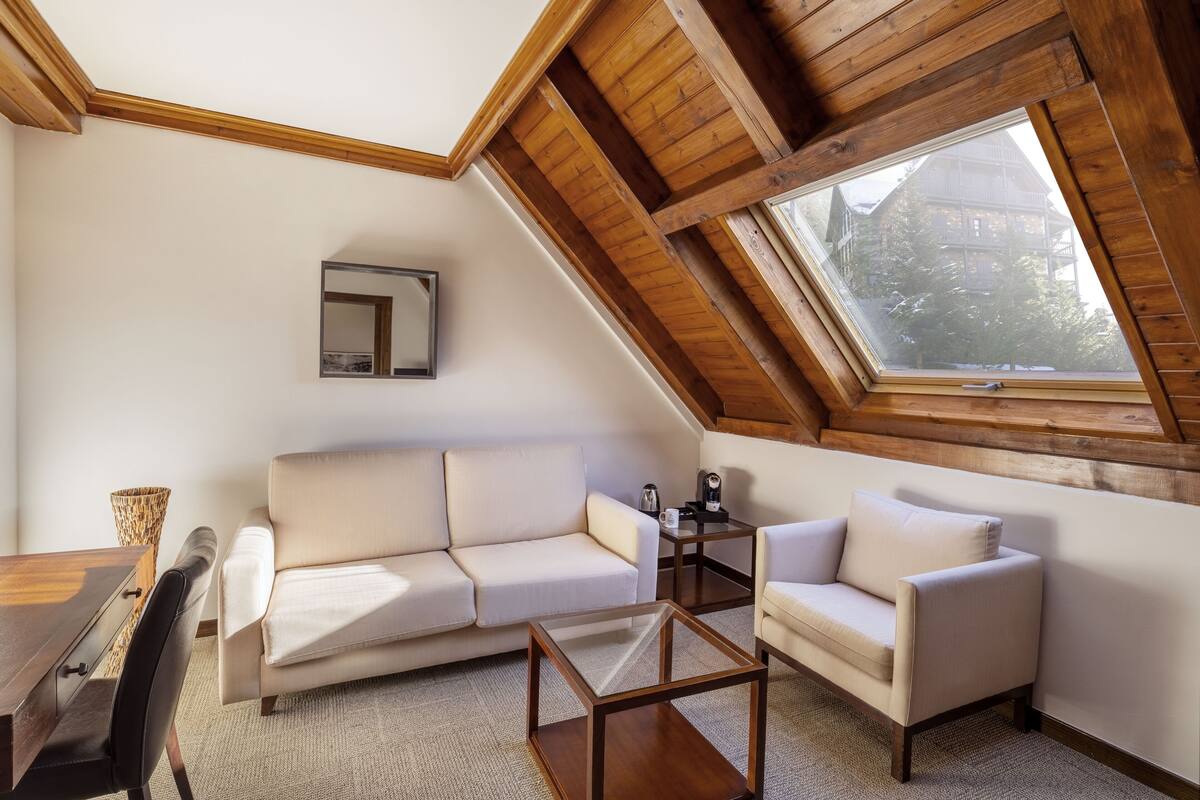 Une chambre mansardée chaleureuse, avec un plafond en pente, une grande fenêtre de toit, un canapé, un fauteuil, une petite table et un miroir.