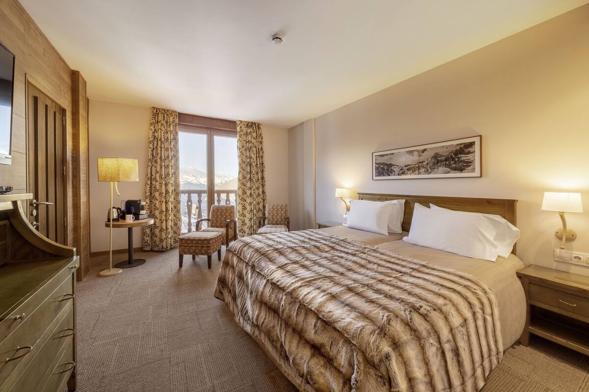 Une chambre d’hôtel avec un grand lit, un bureau avec une lampe, une chaise, une fenêtre avec des rideaux et un tableau encadré au mur.