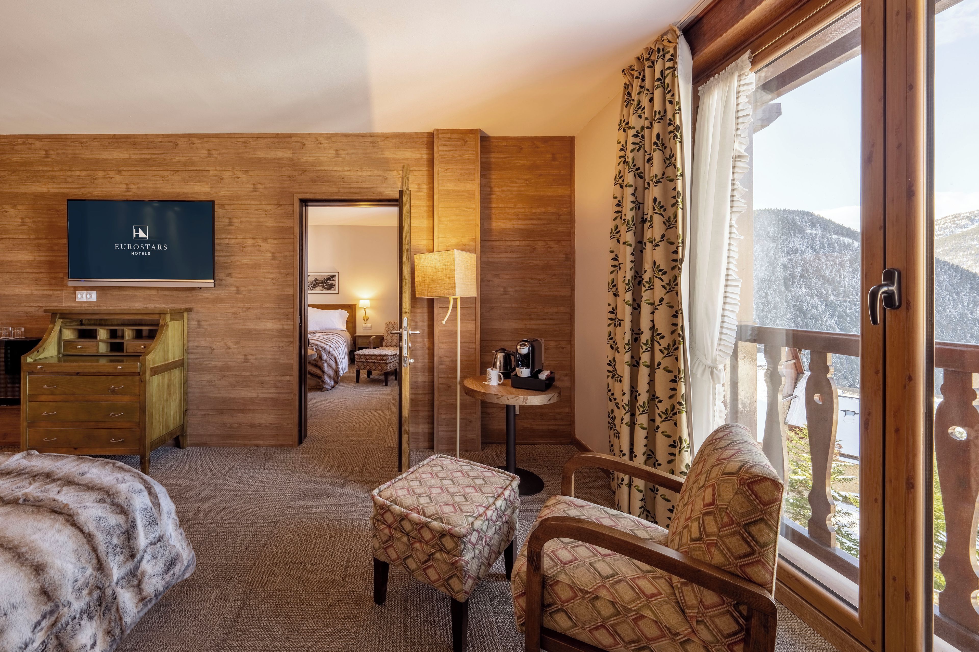 A hotel room with a bed, a chair, a desk, a TV, and a window with a view of snow-covered mountains.