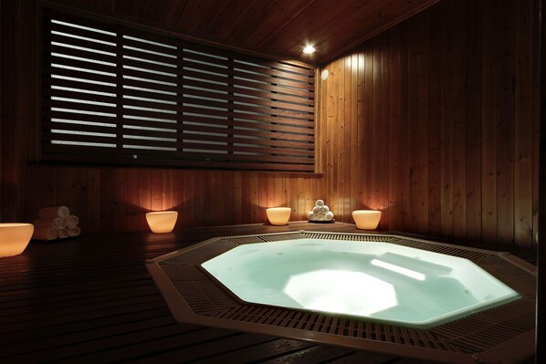 Un sauna de madera con una bañera de hidromasaje central, dos lámparas a los lados y una ventana con persianas horizontales.